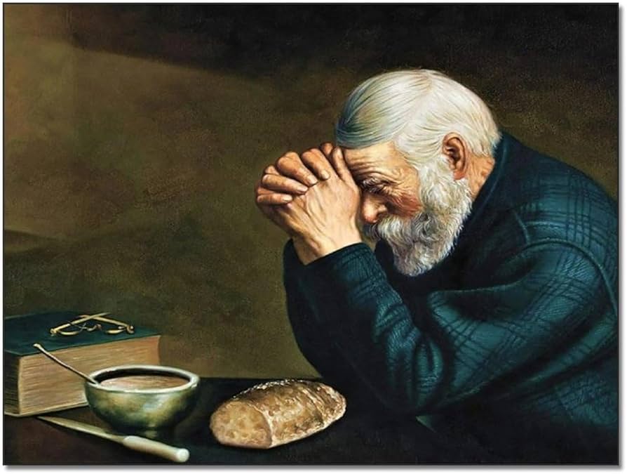 An elderly man with a white beard and short hair, seated at a table with his hands clasped in prayer. In front of him are a bowl of soup, a loaf of bread, and a closed book with glasses on top.